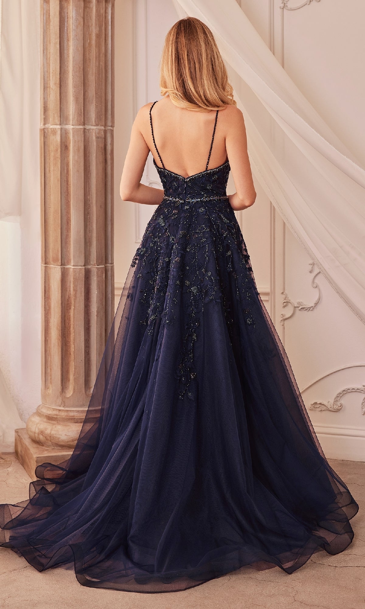 A woman with long blonde hair wears the Long Formal Dress A1251 by Andrea and Leo, featuring a full navy blue tulle skirt, as she stands with her back to the camera in an elegant room.